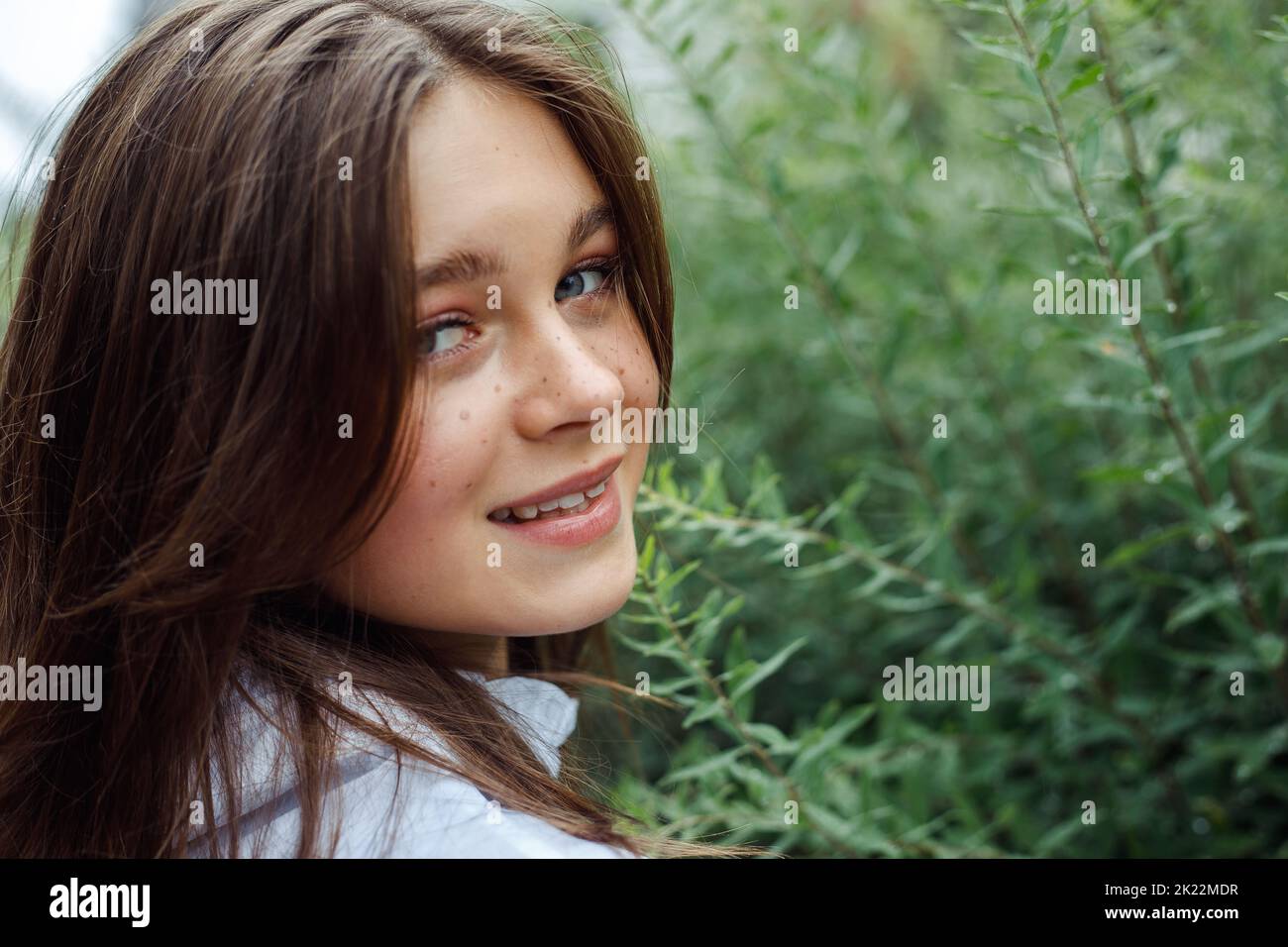 Portrait closeup of charming young girl with stylish makeup and freckles, smiling stand among branches of bushes with green leaves, nature background Stock Photo