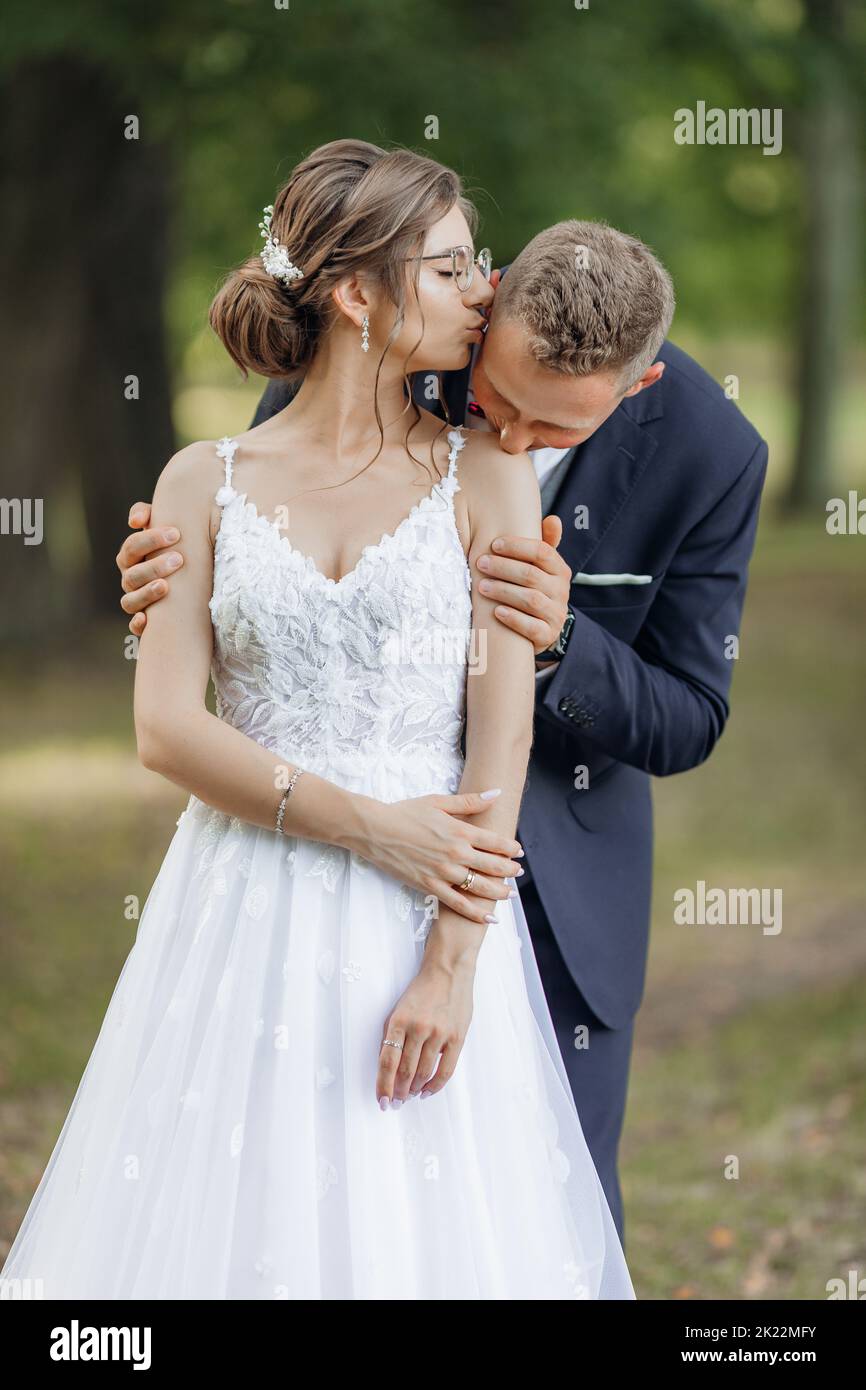 Portrait of amazing wedding couple standing in park in summer. Young beautiful woman bride kissing temple of man. Love. Stock Photo