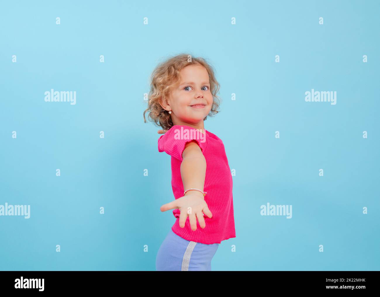 Little smiling, playful, fashionable curly blonde girl in pink shirt and bracelet show hand to camera. Dancing activity Stock Photo