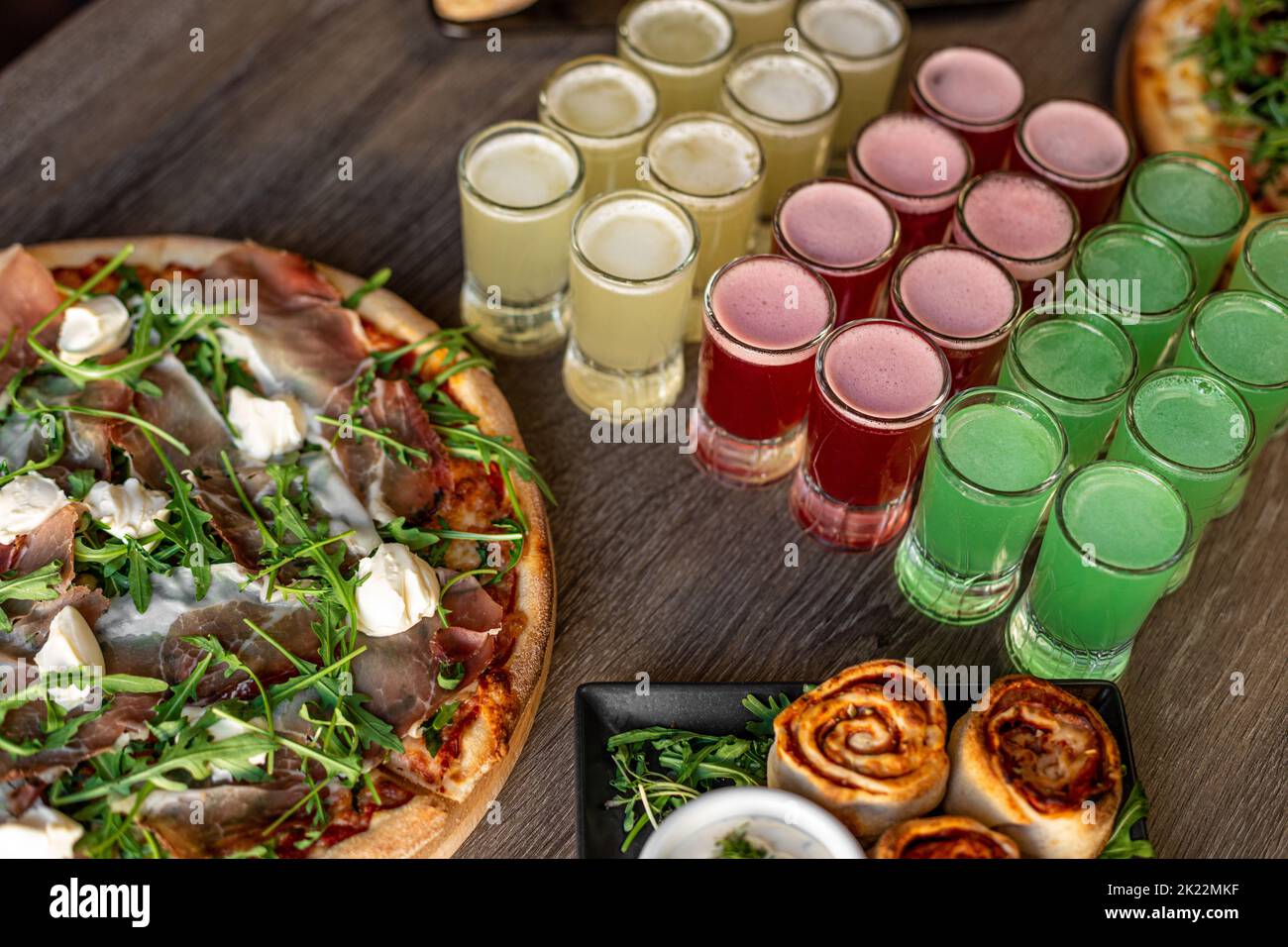 Pizza with mozzarella cheese, bacon, meat on cafe table. Alcohol colorful red, yellow and green jelly drinks and rolls Stock Photo
