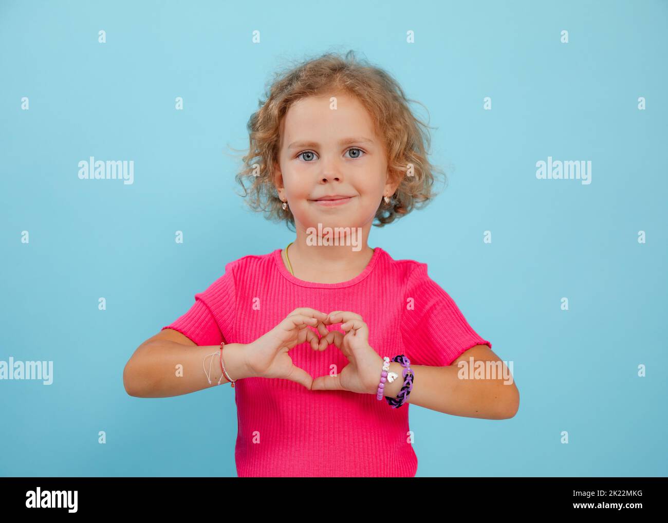 Small expressive, confident, playful, positive curly blonde girl showing heart sign by hands. Love and care. Copy space Stock Photo