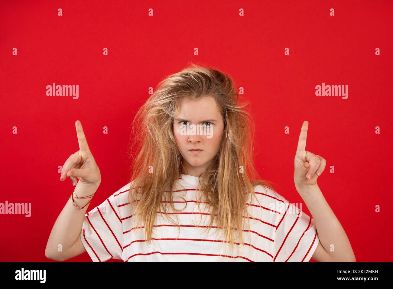 Puzzled, shocked blonde teenage girl in striped t shirt showing by index fingers empty space above head. Hot news, ad Stock Photo