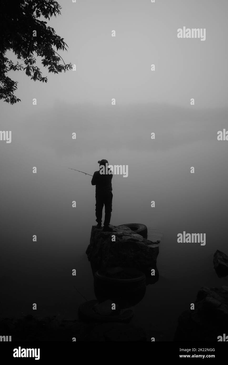 A greyscale shot of man standing on a rock in the middle of the lake Stock Photo