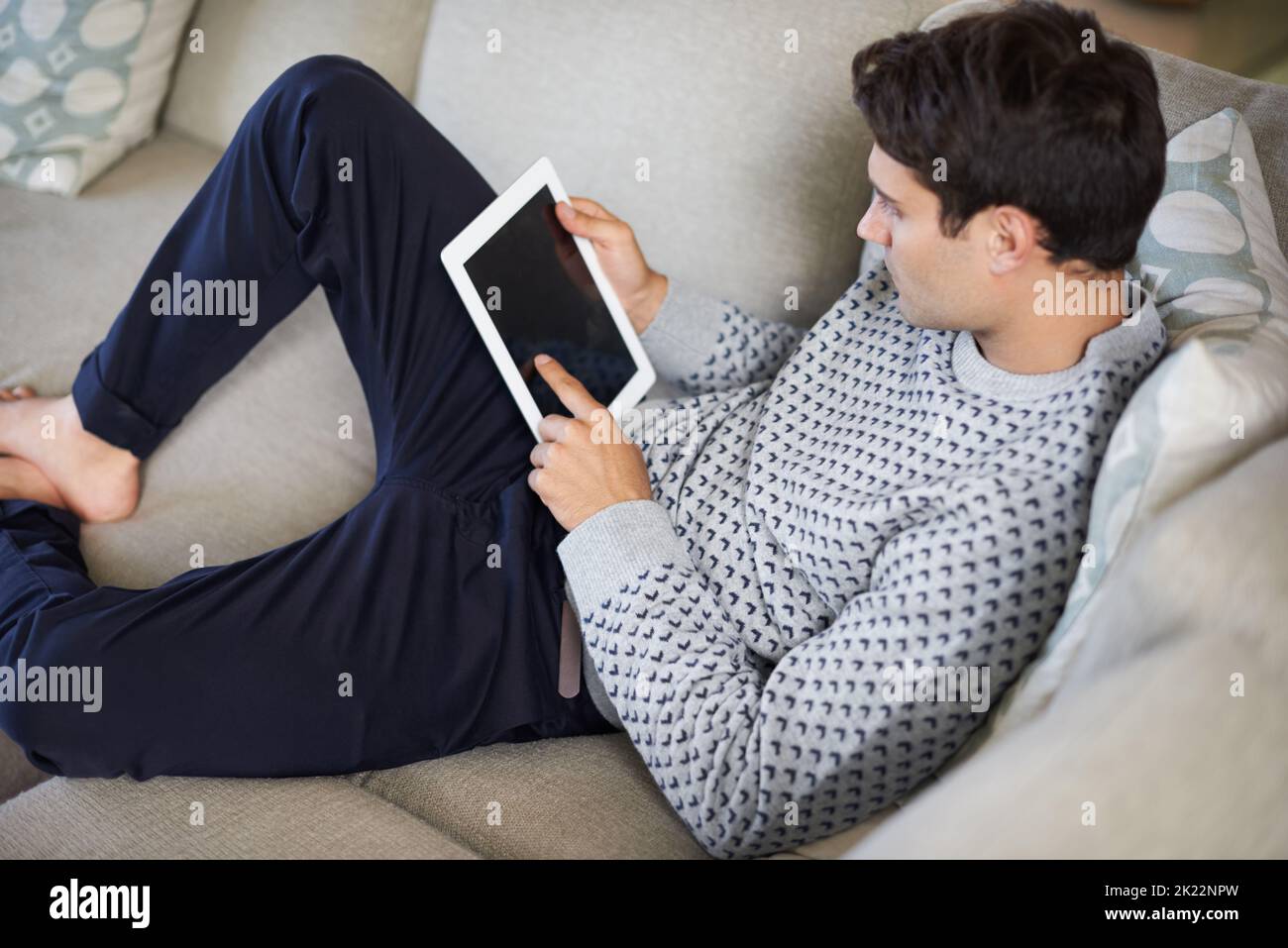 Where do i switch this thing on. a young man lying on his sofa while using his digital tablet. Stock Photo
