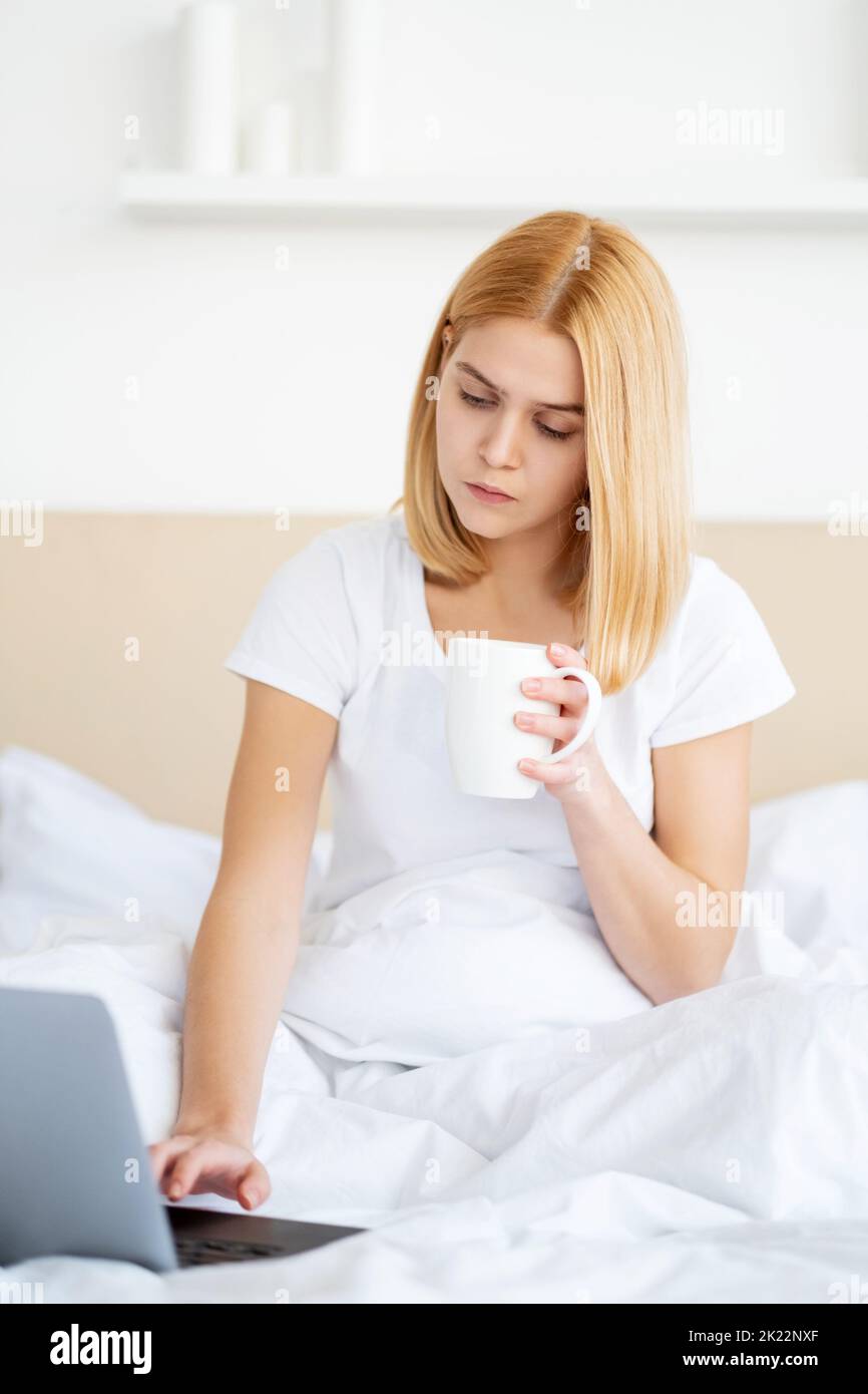Work from home. Remote job. Freelance lifestyle. Bedtime leisure. Busy young woman employee internet browsing on laptop in bed with cup of coffee at l Stock Photo