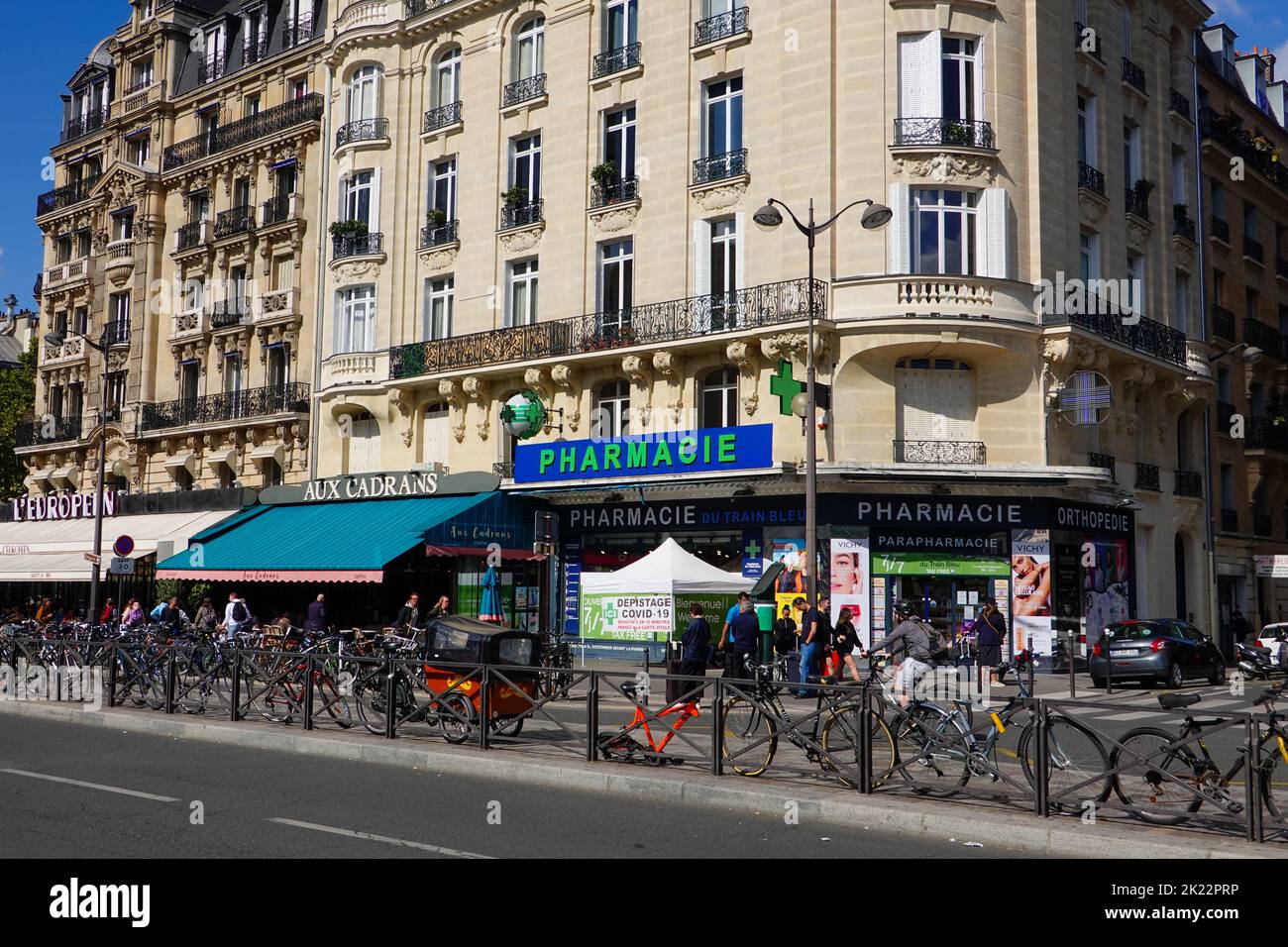 People, bicycles, busy area across from a Paris train station with a pharmacy, offering Covid testing, and other shops located in Haussmann building. Stock Photo