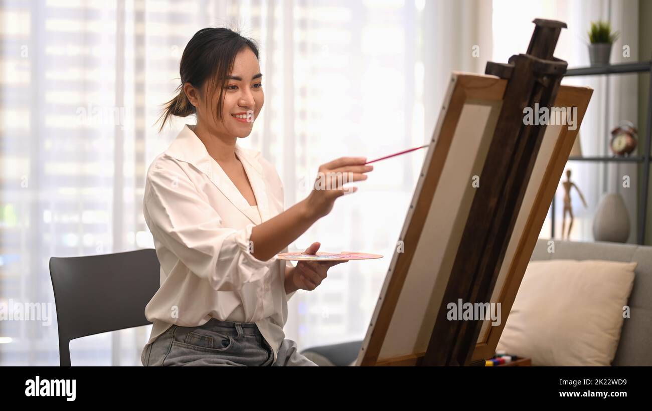 Happy young female artist holding palette and painting picture on ...