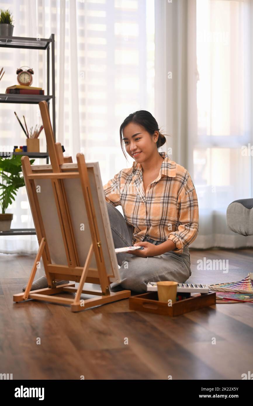 Pleasant young woman in casual wears sitting on floor in front of canvas and painting with water color Stock Photo