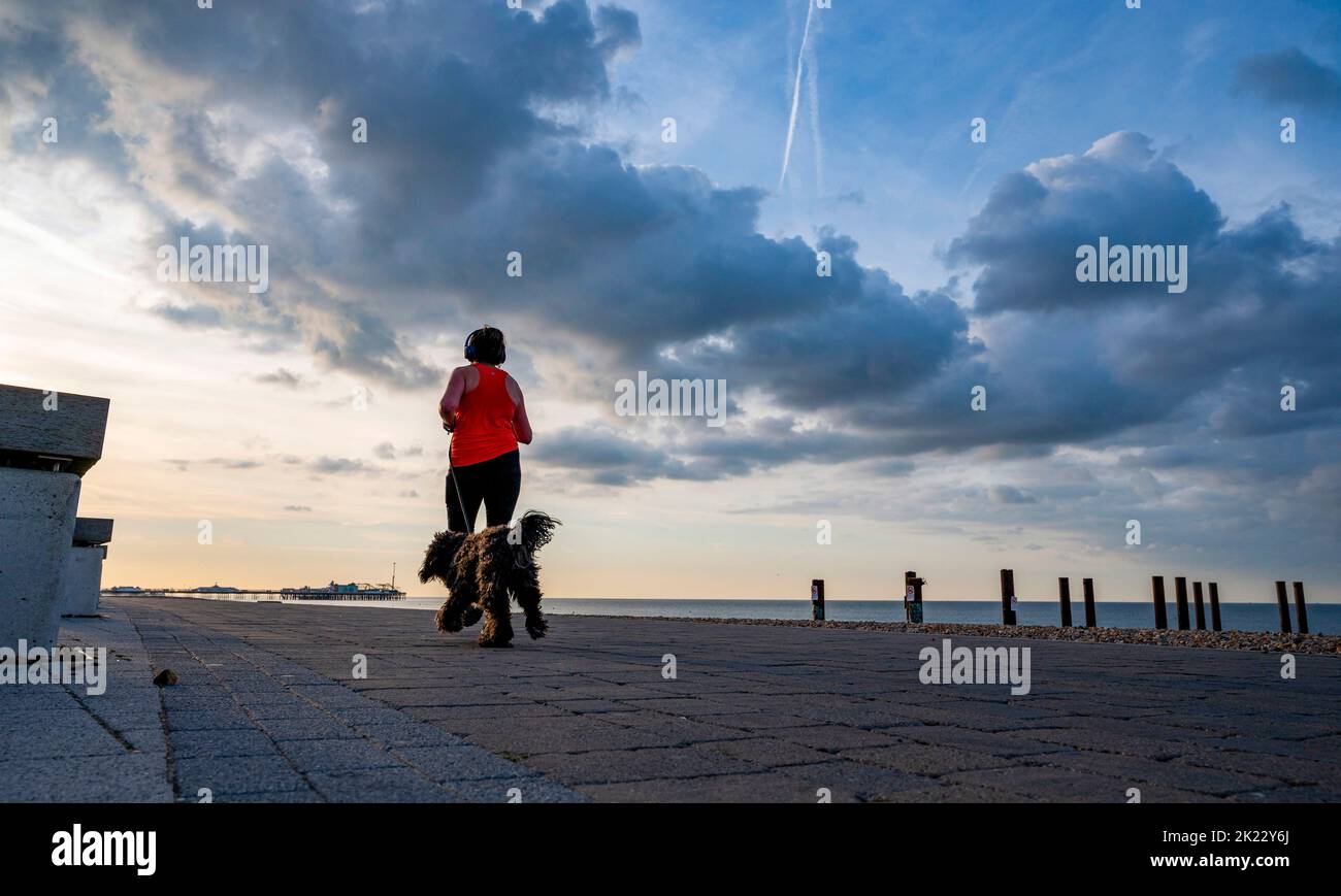 Brighton UK 22nd September 2022 - A runner and their dog pass under  dark clouds and early morning sunshine over Brighton seafront as more unsettled weather is forecast for the next few days .   . : Credit Simon Dack / Alamy Live News Stock Photo