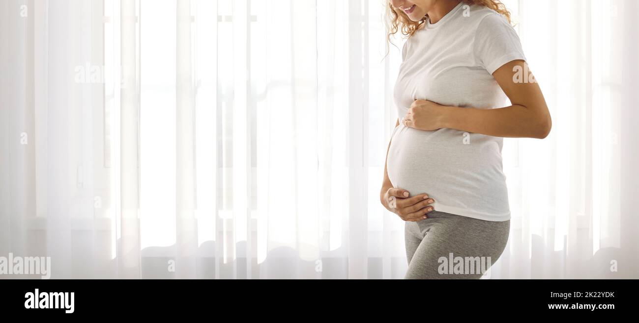 Expectant mother gently strokes and hugs her pregnant tummy while standing against bright window. Stock Photo