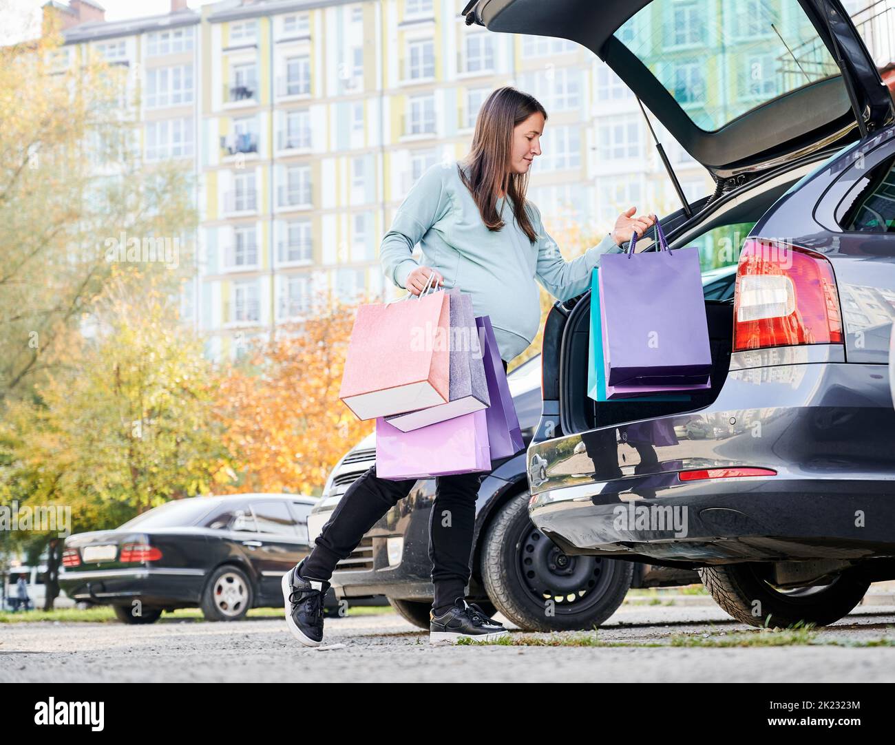 Caucasian woman with belly puting shopping bags in vehicle trunk enjoying maternity and parenthood in awaiting mode. Pregnant female with purchase preparing for baby birth during weekend in city. Stock Photo