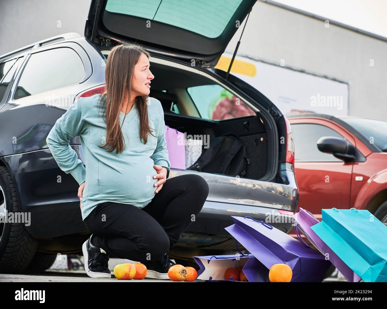 Pregnant woman waiting for ambulance during premature childbirth at car parking in city. Female with paper bags feeling pain in belly abdomen have birth contractions after shopping time. Stock Photo