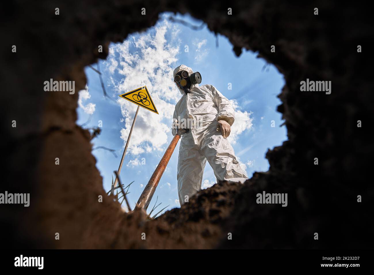 Below view on male ecologist digging pit by shovel, wearing protective overalls and gas respirator near biohazard symbol warning about dangerous biological materials. View from inside pit. Stock Photo