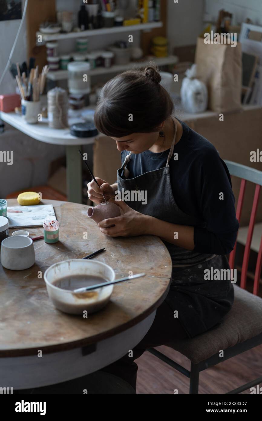 Lady master paints clay jug with serious and concentrated expression sitting at table in workshop Stock Photo