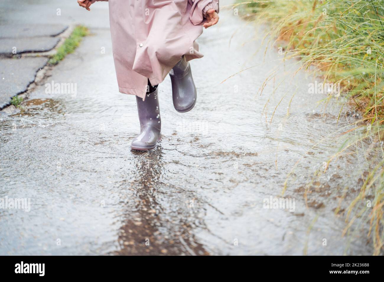 Little girl in pink waterproof raincoat, purple rubber boots funny jumps through puddles on street road in rainy day weather. Spring, autumn. Children Stock Photo