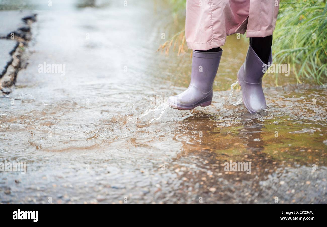 Little girl in pink waterproof raincoat, purple rubber boots funny jumps through puddles on street road in rainy day weather. Spring, autumn. Children Stock Photo