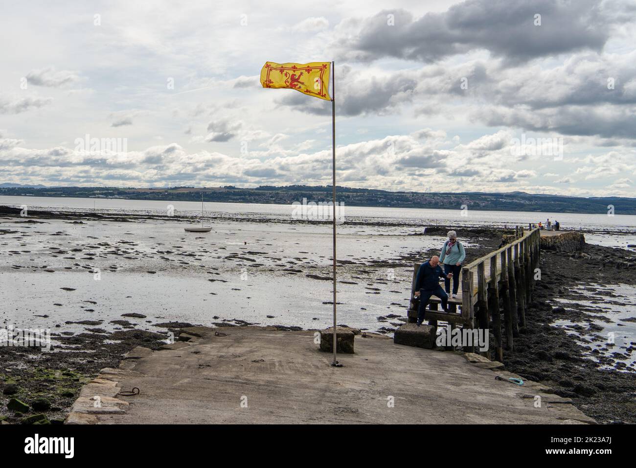 The Royal Banner of Scotland flag flying at a pier in the Firth of Forth in Culross, Scotland, UK, with two people descending from the pier. Stock Photo
