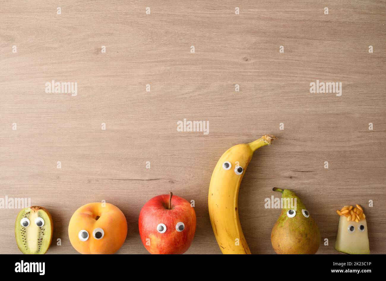 Background of row of funny fruit with eyes on wooden bench as a concept for motivating children to eat. Top view. Stock Photo