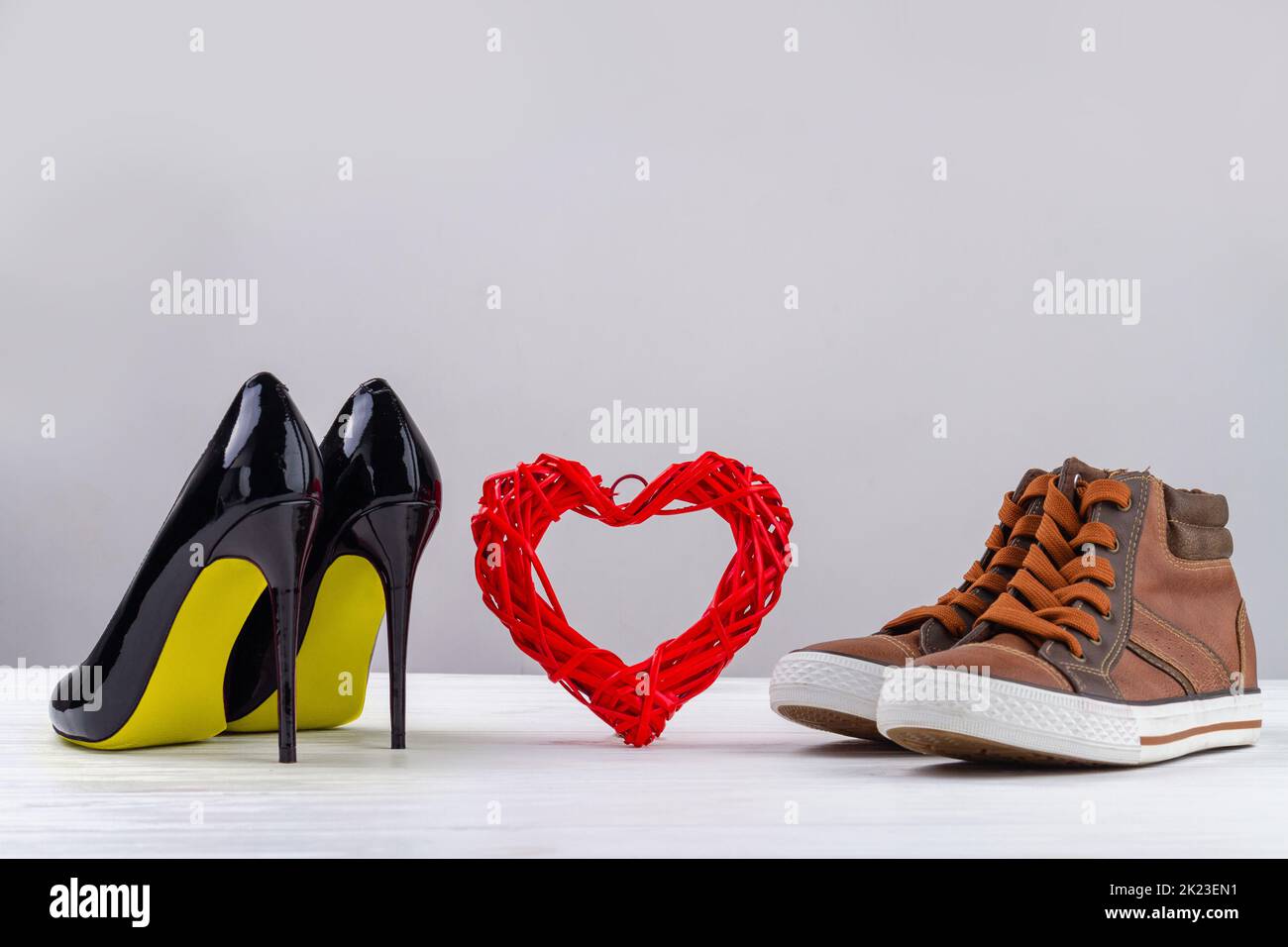 Red knitted heart with black womens high heel shoes and mens sneakers. Footwear on white background with copy space. Stock Photo