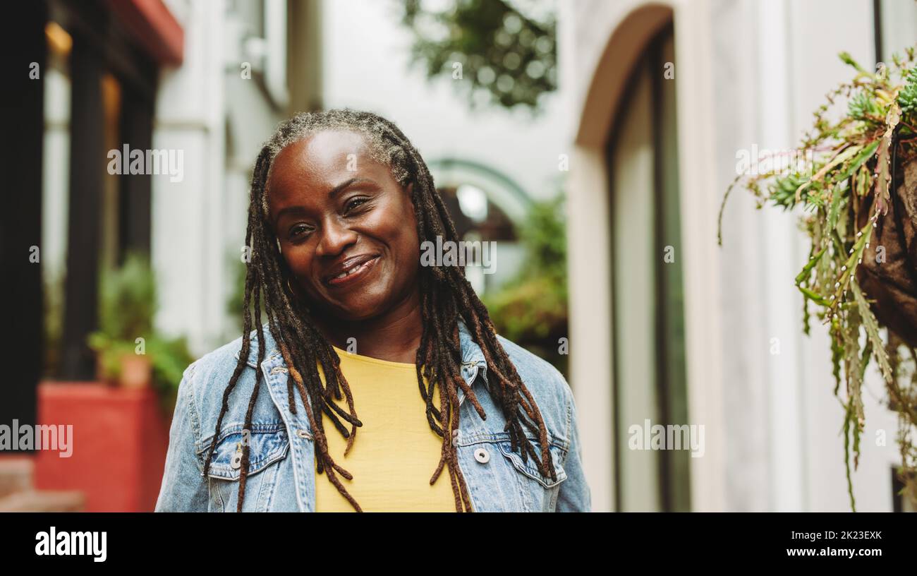 Woman with dreadlocks smiling happily while standing outdoors. Cheerful senior woman going out in casual clothing. Stock Photo