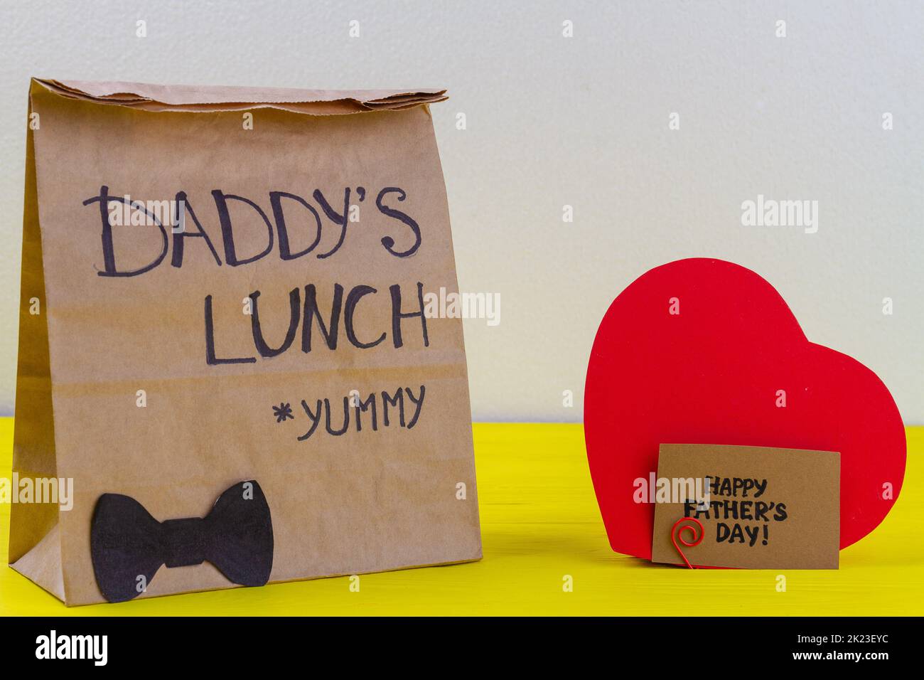 Paper bag and red heart on yellow wooden desk. Daddys lunch for fathers day. Stock Photo