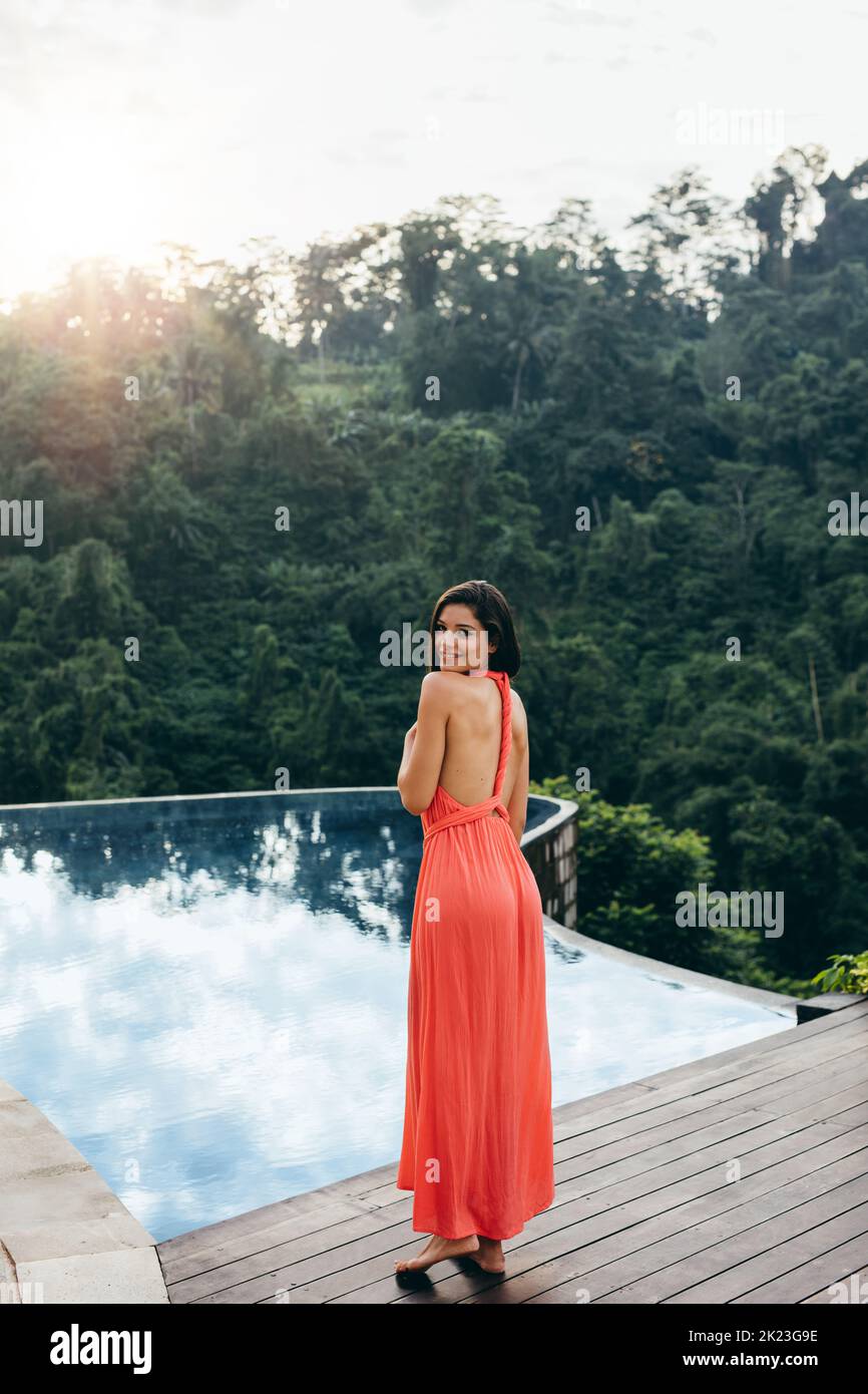 Full length portrait of beautiful young woman standing at poolside and looking over shoulder. Caucasian female model in sundress near swimming pool of Stock Photo