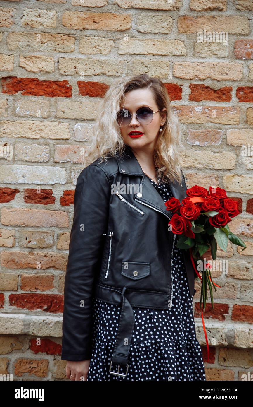 Portrait of young woman with long wavy fair hair, red lipstick holding bouquet of red roses, standing at brick wall. Stock Photo