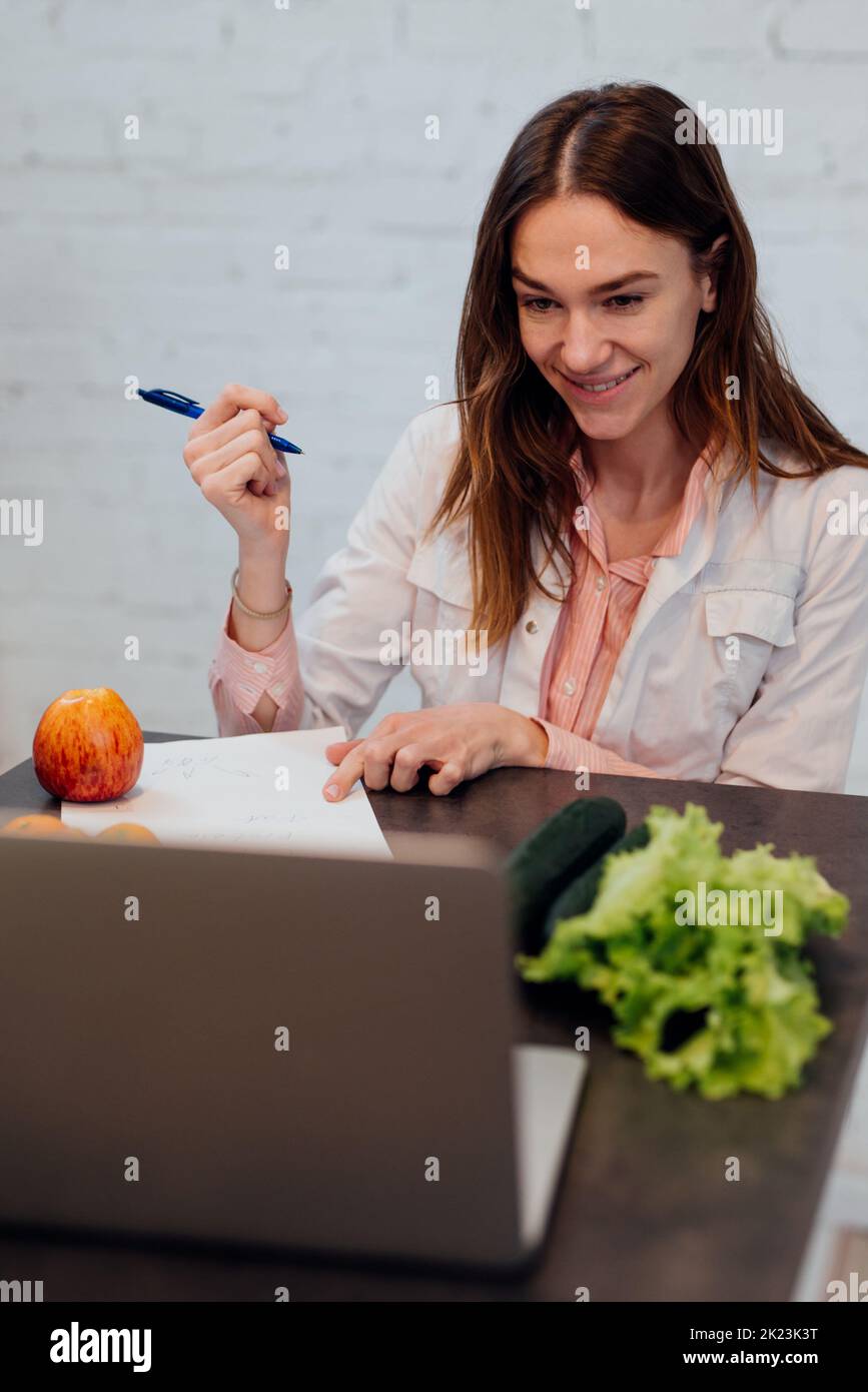 Physician nutritionist consults online. A female doctor sits at her desk in front of her laptop and talks on a video call. Stock Photo