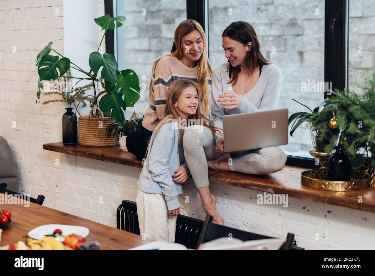 Female friends and a little girl sitting at home on a windowsill watching a movie on a laptop Stock Photo