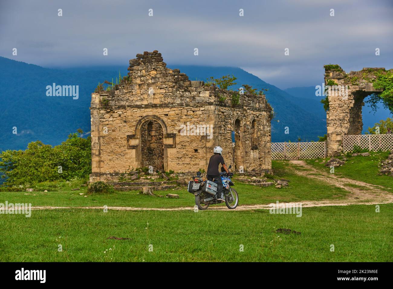 A man on a motorcycle rides along a dirt road among green grass to the ruins of an ancient Bedia temple. Stock Photo