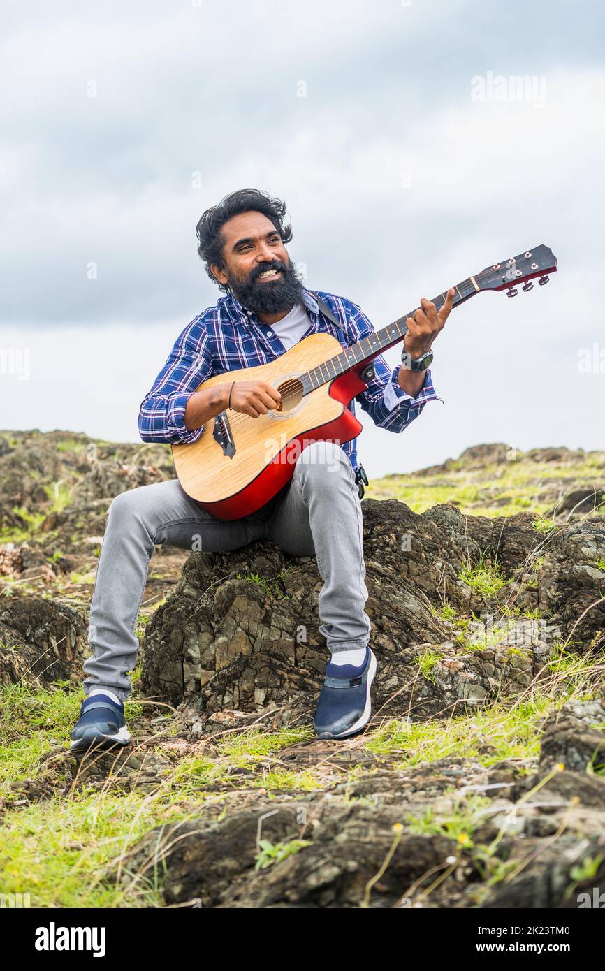 vertical shot of Young beard man playing guitar by singing song while sitting on top of hill - concept of hobbies, leisure activity and entertainment Stock Photo