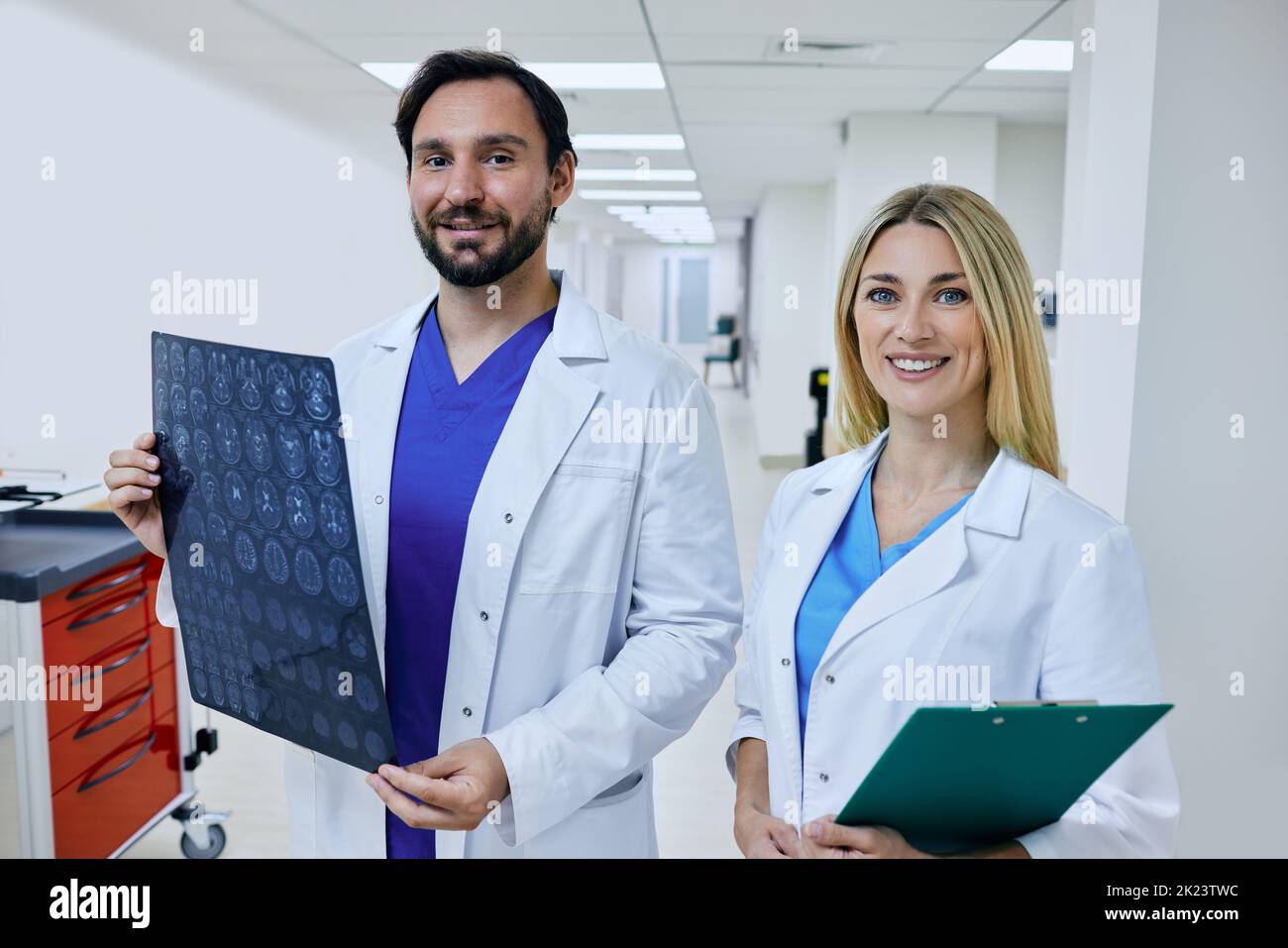 Radiographer with MRI of patient head is standing in corridor of hospital with female radiologic technologist and at looking at camera. Team working i Stock Photo