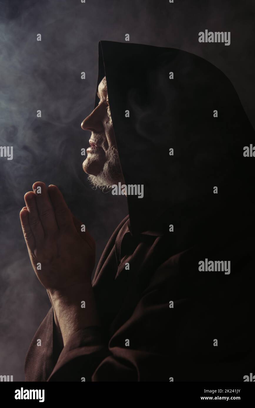 side view of medieval monk in hooded robe praying on black with smoke ...