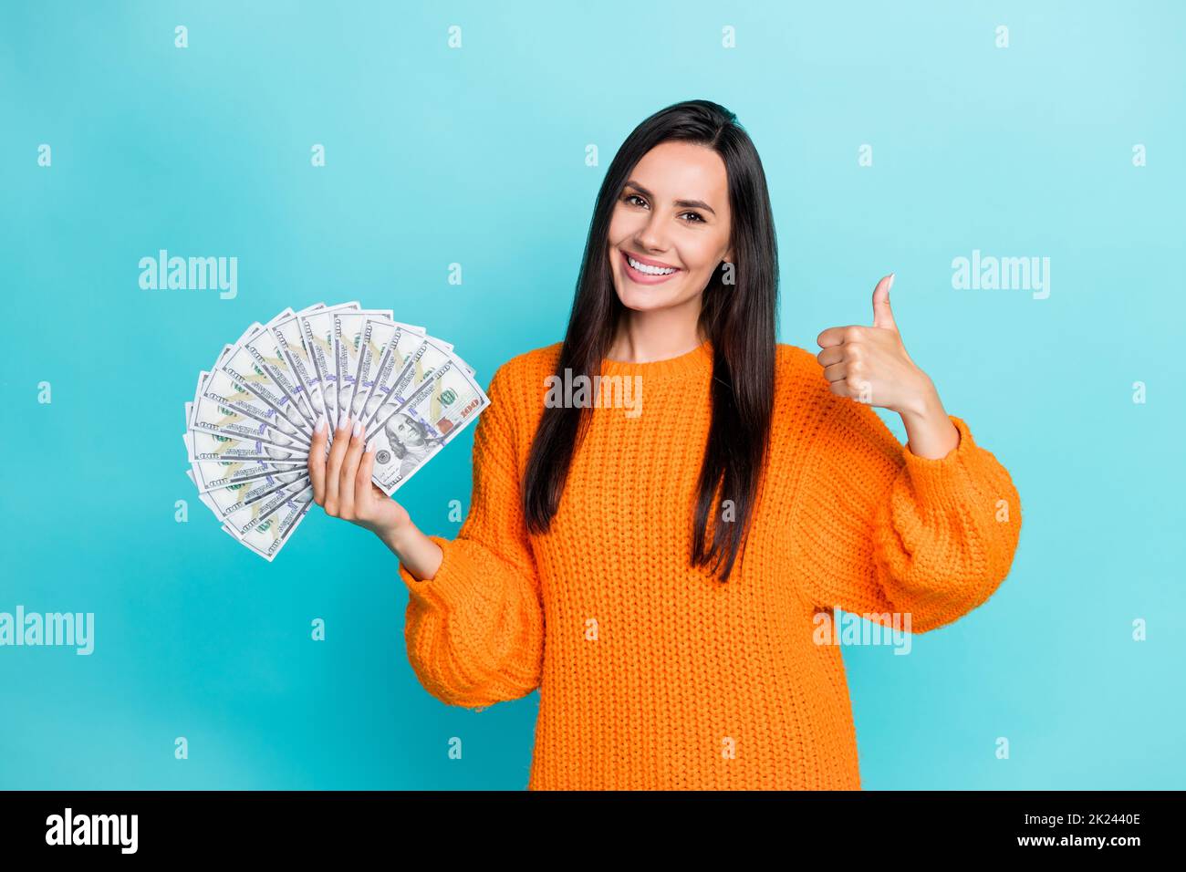 Photo of adorable cute girl with straight hairdo dressed orange sweater showing thumb up holding money isolated on turquoise background Stock Photo