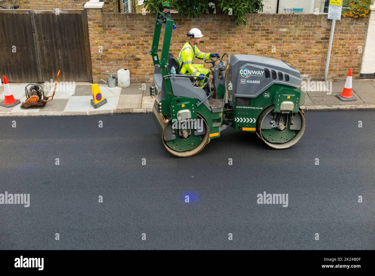 Road roller smoothing hot tarmac that has been laid well UK while resurfacing of a residential Street in Twickenham, Greater London, UK. The previous worn and potholed surface has been removed. The blue light shining on the ground is the blue light exclusion zone for safety reasons, operated with heavy machinery such as this compactor. (132) Stock Photo