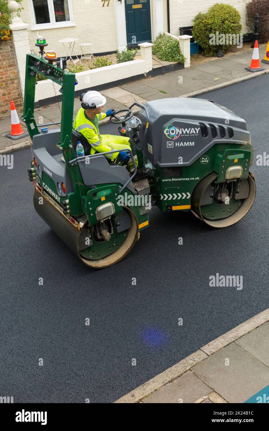 Road roller smoothing hot tarmac that has been laid well UK while resurfacing of a residential Street in Twickenham, Greater London, UK. The previous worn and potholed surface has been removed. The blue light shining on the ground is the blue light exclusion zone for safety reasons, operated with heavy machinery such as this compactor. (132) Stock Photo