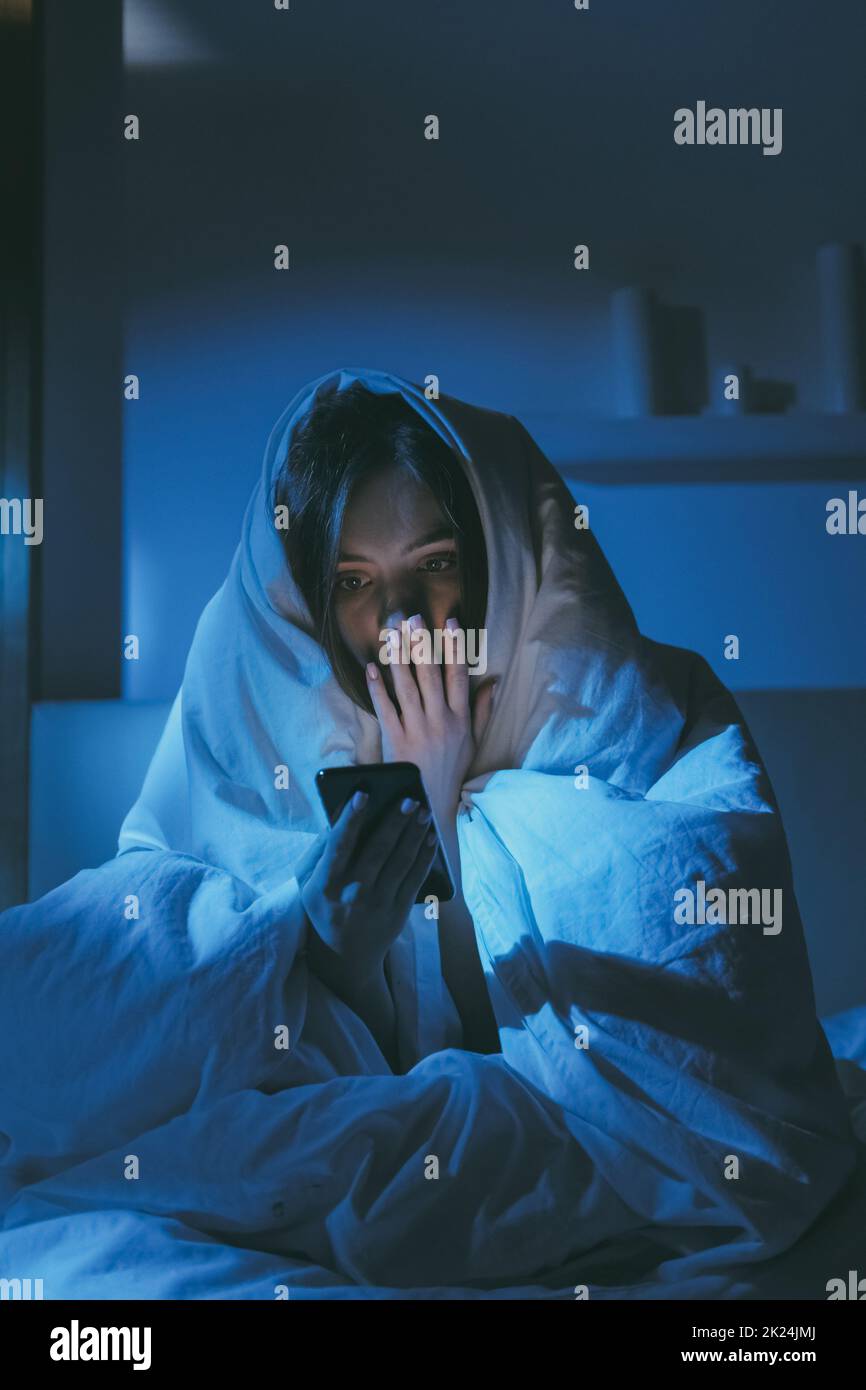 Night insomnia. Late online. Stress anxiety. Social media addiction. Terrified scared girl reading disturbing Internet news on mobile phone sitting on Stock Photo