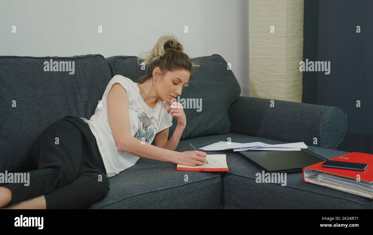 Pretty woman makes notes in notebook while lying on couch at home in a modern apartment. Is preparing for important meeting. Very focused prepares the Stock Photo