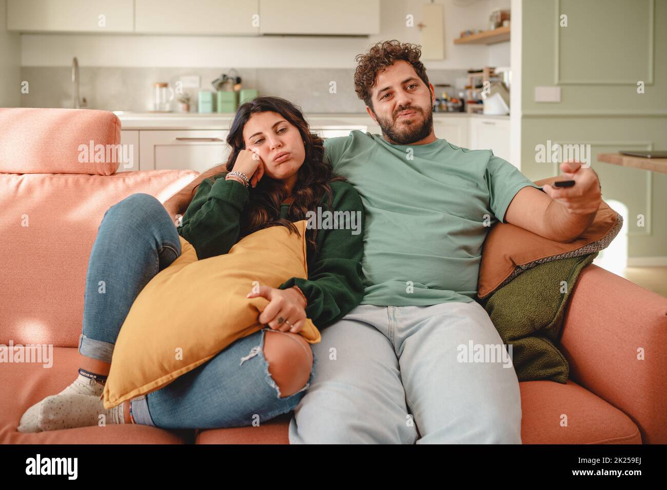 Bored couple watching tv sitting on the sofa - newlyweds cuddling together - the man holding the remote control switches channels by zapping - people Stock Photo