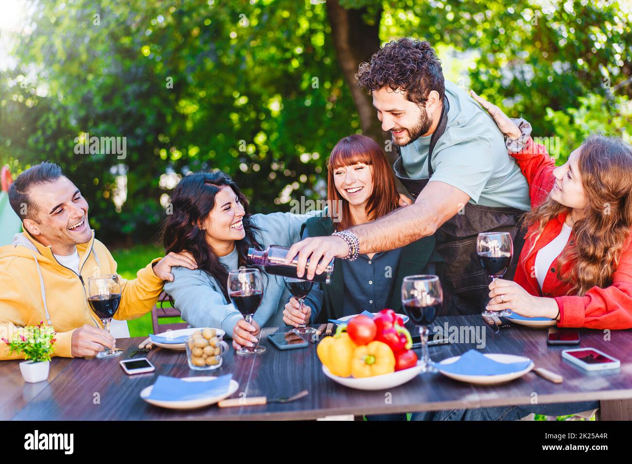 Man pouring red wine into glass while enjoying dinner party with friends outdoors in the backyard - happy friends having fun drinking wine in the terr Stock Photo