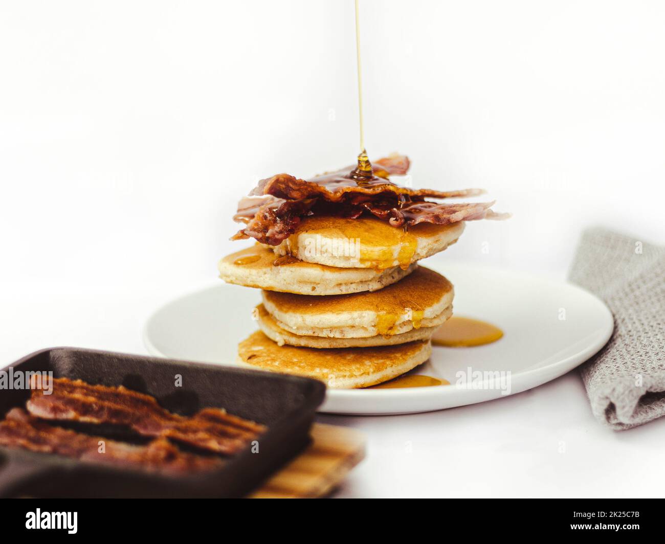 Maple Syrup being poured on to pancake stack topped with crispy bacon.  Cast-iron skillet in the foreground. Stock Photo