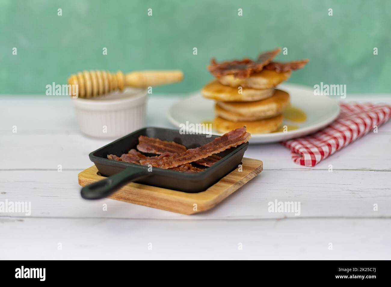 Mini cast-iron skillet with crispy bacon and pancake stack with  honey topping in the background Stock Photo