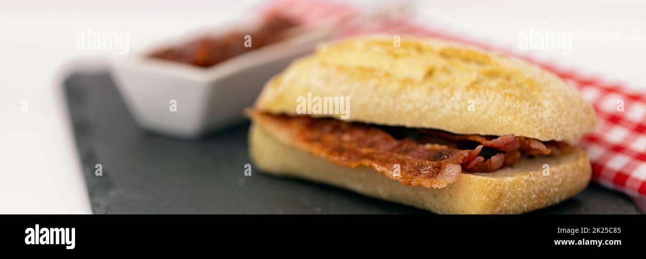 Panorama of crispy bacon in a sourdough roll with tomato relish on a slate with gingham napkin Stock Photo