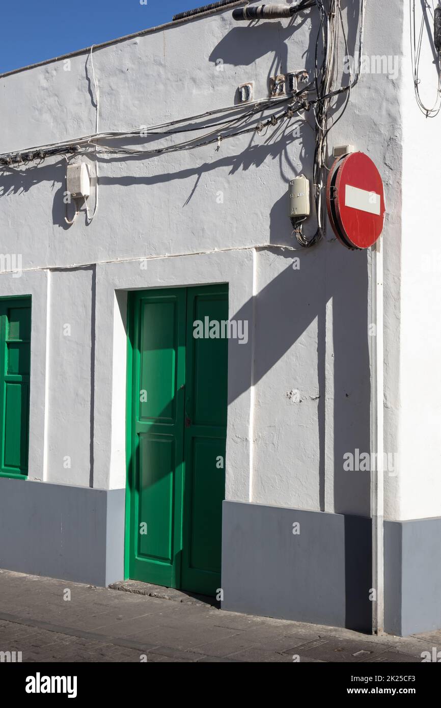 Corner of a low building, with a typical white facade and green door and window shutter. Many cables on the wall. No entry traffic sign. Brigt blue sk Stock Photo