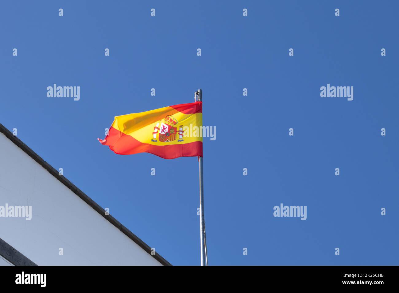 Triangle of a roof of a building and a pole with spanish flag, blowing in the wind. Bright blue sky. Arrecife, Lanzarote, Canary Islands, Spain. Stock Photo