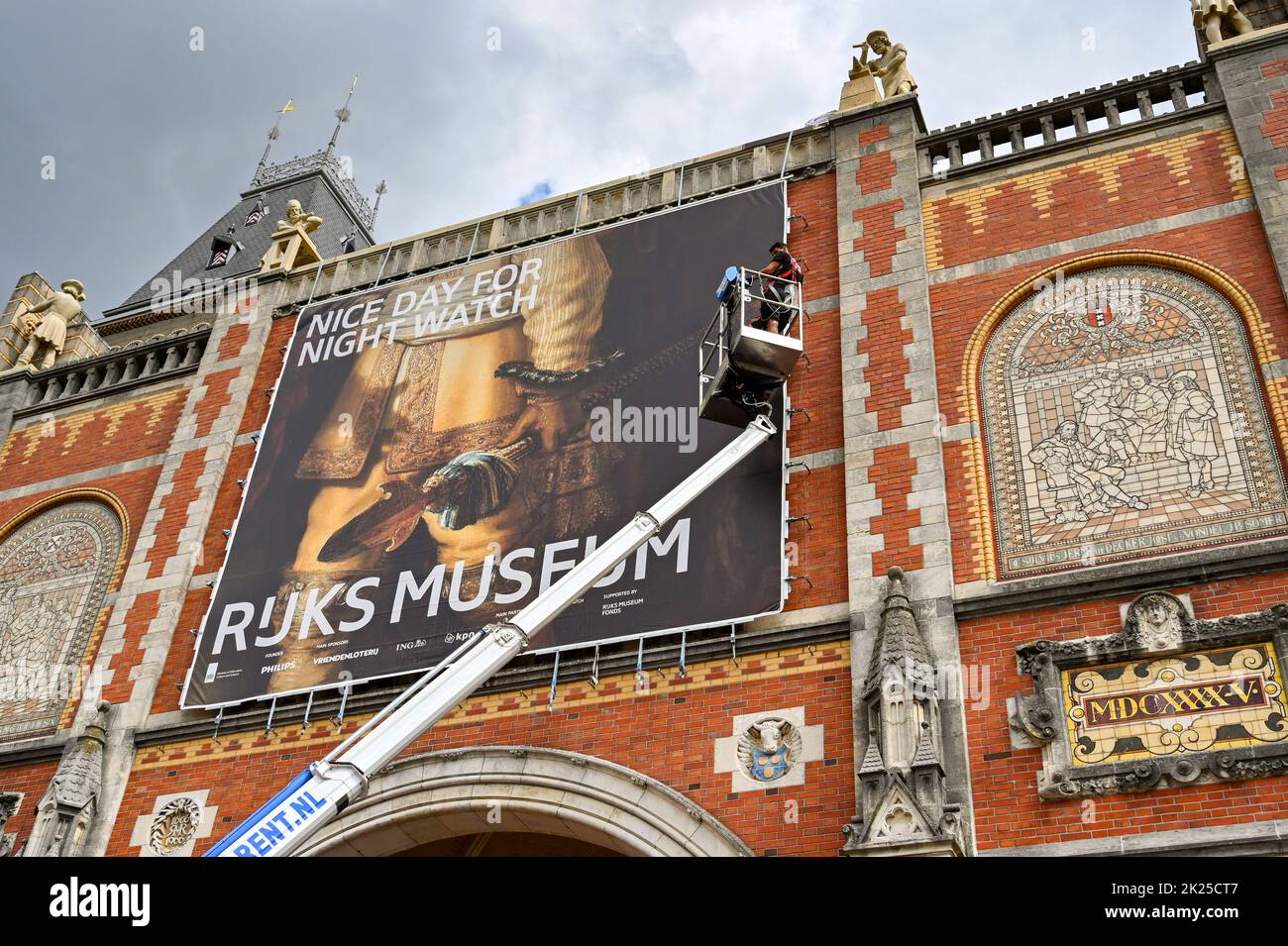Amsterdam, Netherlands - August 2022: People standing on a cherry picker platform fixing a large banner to the outside wall of the famous Rijks Museum Stock Photo