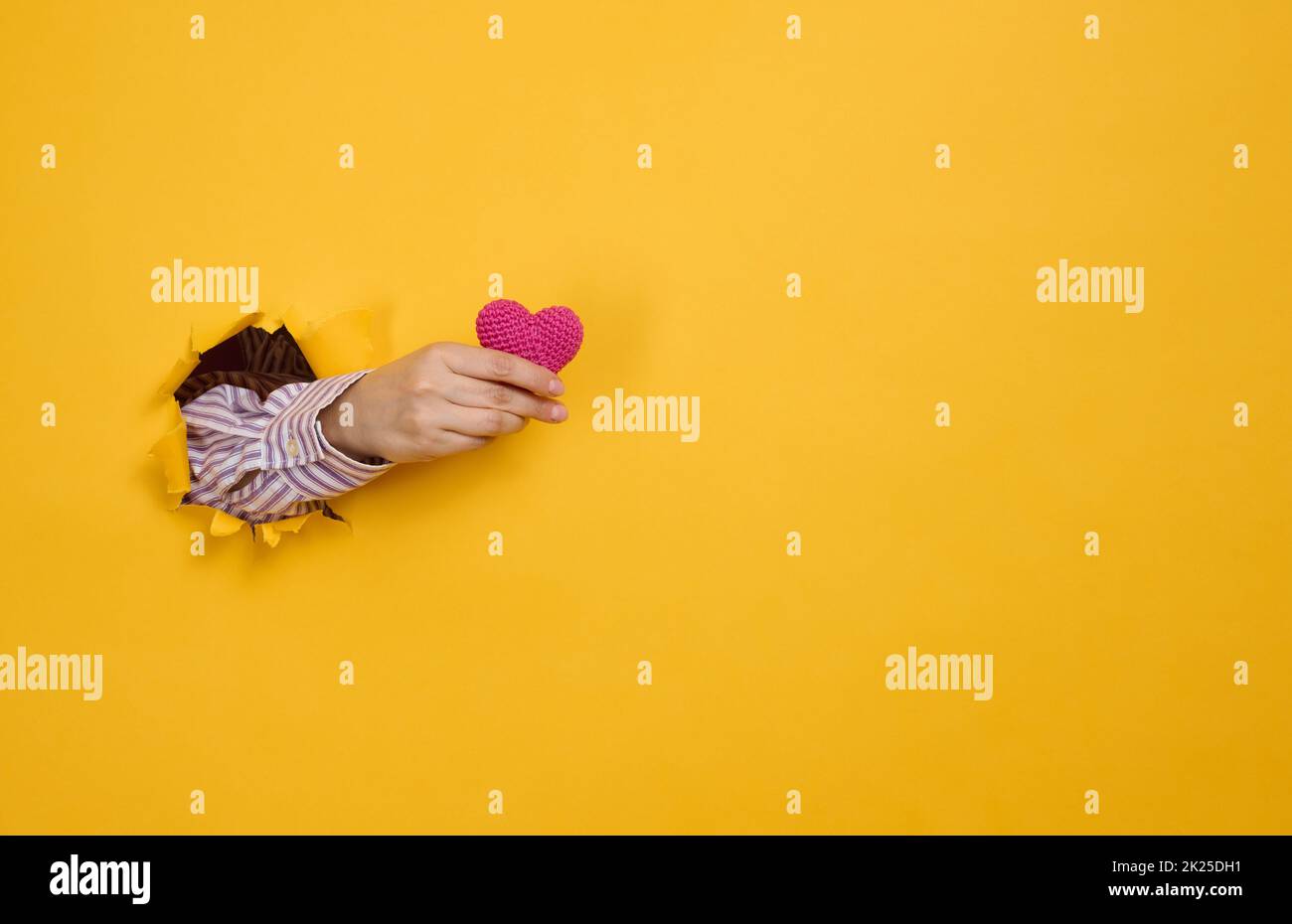 female hand holding a red heart on a yellow background. Part of the body sticks out of the torn paper Stock Photo