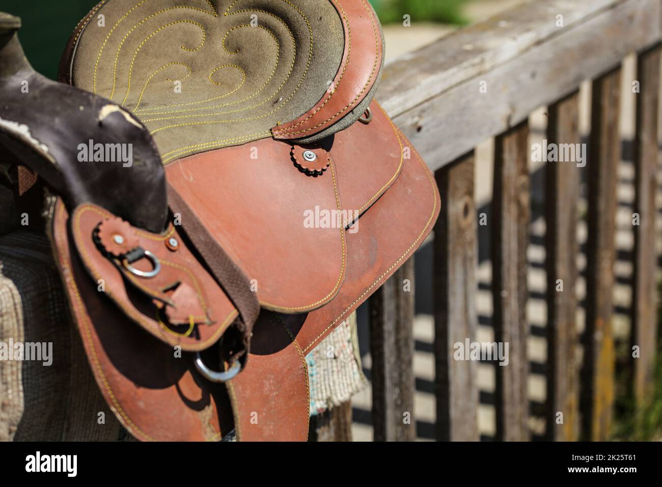 Old worn leather horse saddle on wooden fence, lit by sun Stock Photo