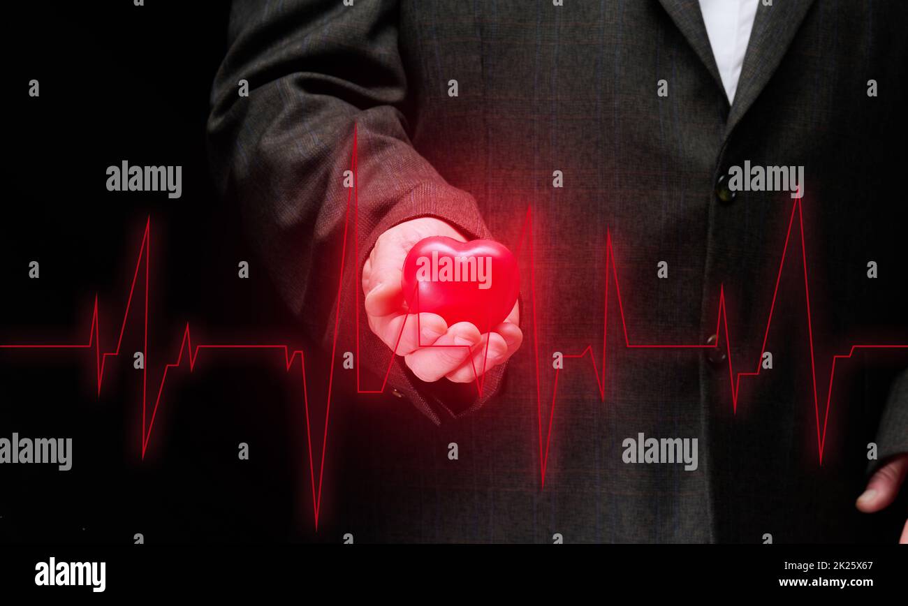 businessman holding a red heart on a black background, in front of him is a cardiogram. Office workers cardiovascular disease concept Stock Photo