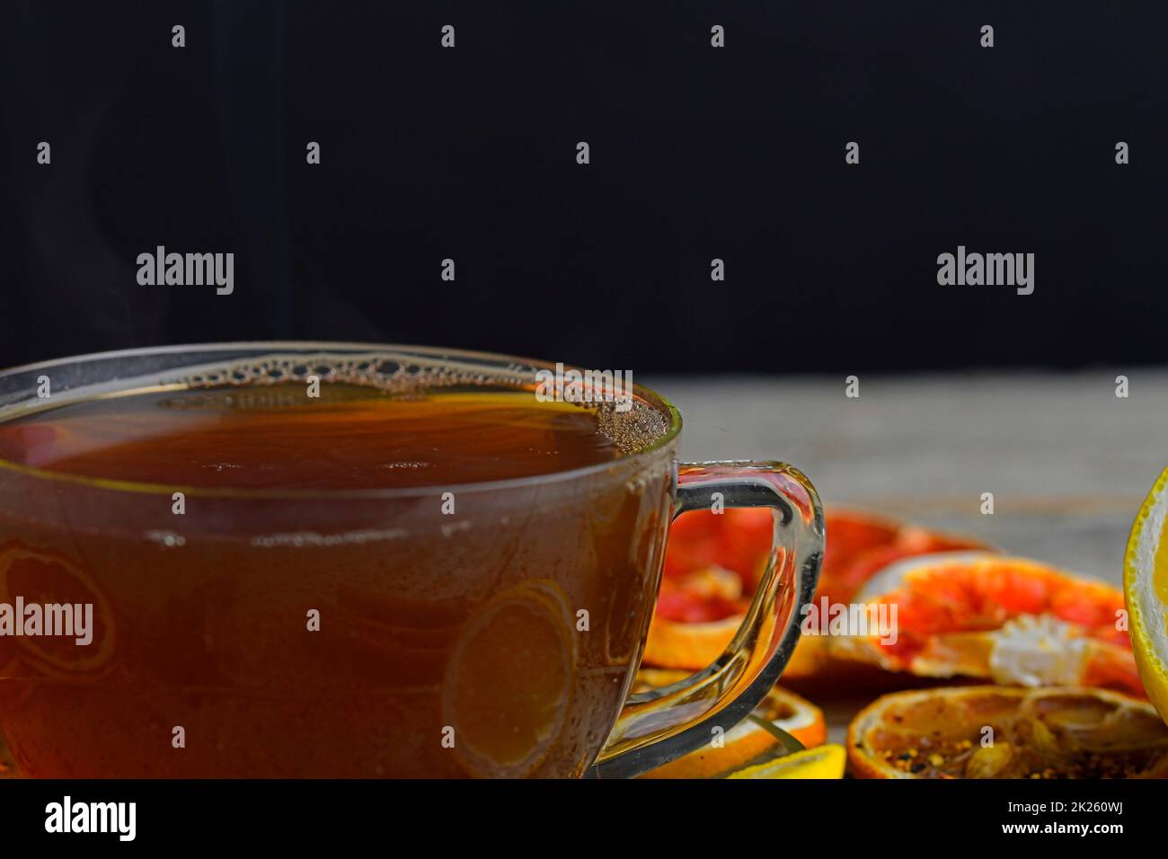 Cup with hot tea and steam on black. Glass cup of black tea with cinnamon sticks, anise star, lemon and dried fruit on white wooden table background. Macro image Stock Photo